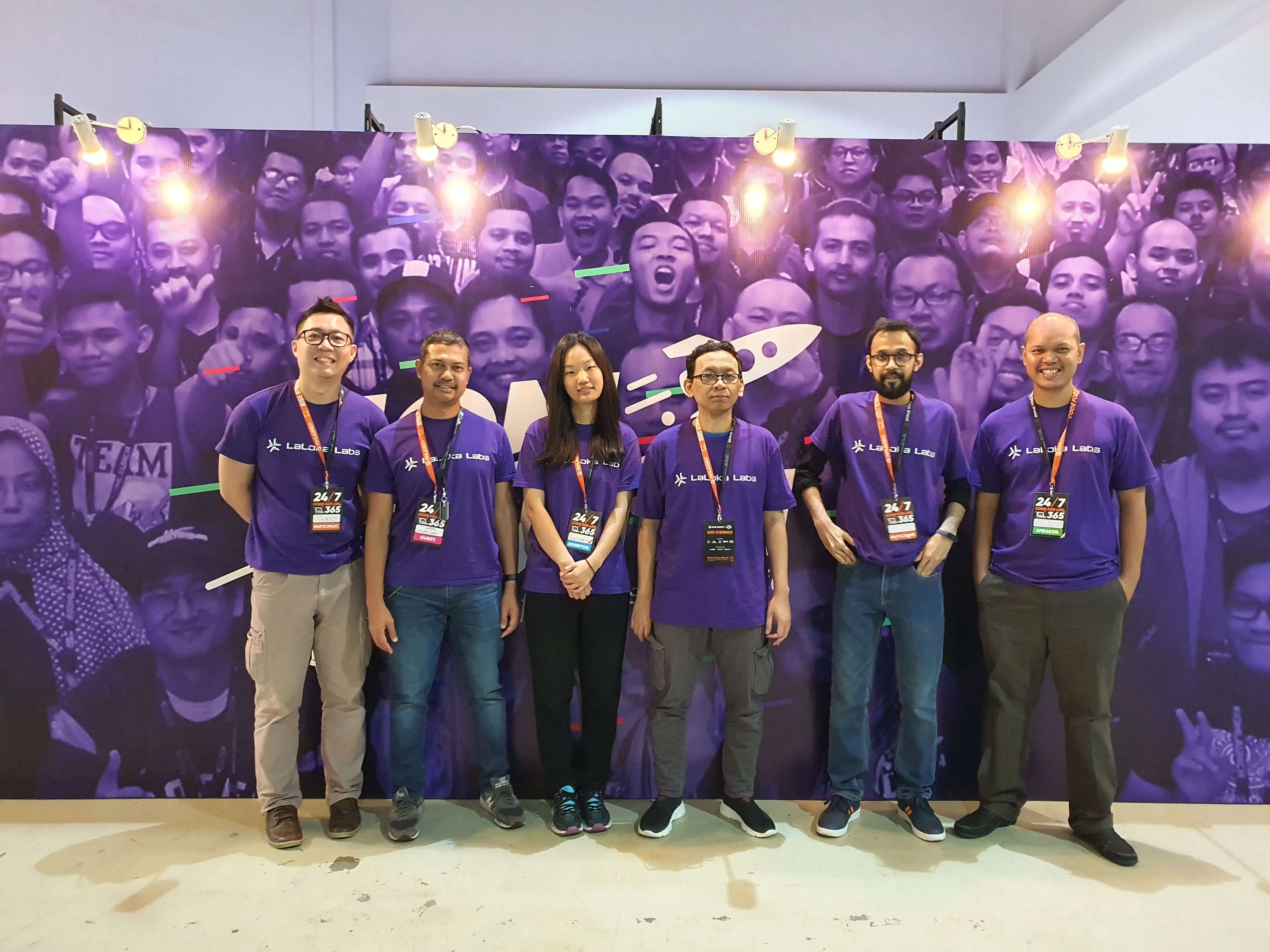 Group photo: The Kafkai (LaLoka Labs) team of people posing in purple shirts in front of a large purple branded backdrop with faces in the background.